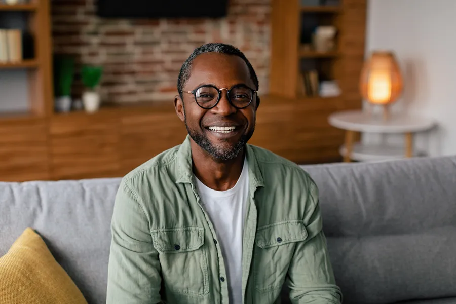 A middle aged black man in a green button up shirt sits on his couch smiling, happy with his treatment for andropause and low testosterone from Carolyn Zaumeyer, MSN, APRN at LowTE Florida® in Greater Fort Lauderdale.