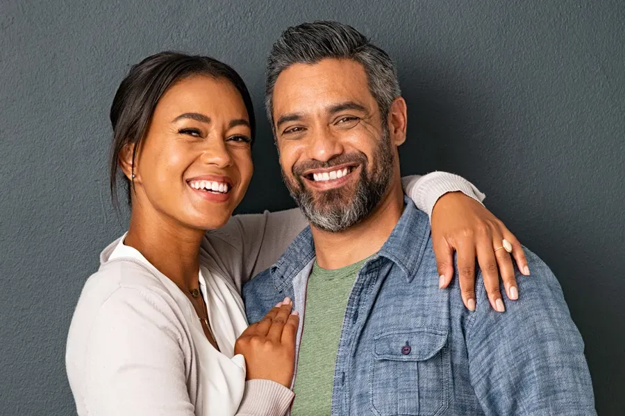 A middle-aged couple with their arms around each other standing against a gray background and smiling, happy with the bioidentical hormone therapy they received from Carolyn Zaumeyer, MSN, APRN in Fort Lauderdale.