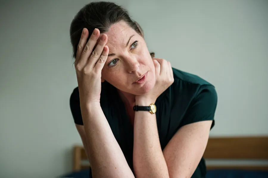 A woman in a dark blue dress sits on her bed holding her head in her hands with an exhausted expression. Get help for chronic fatigue syndrome from Carolyn Zaumeyer, MSN, APRN at LowTE Florida® in [AERA].