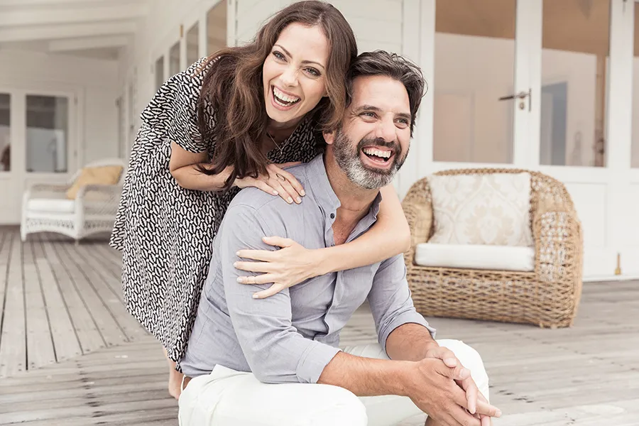 Health-and-Wellness-Services-Doctor A middle-aged couple on a wooden porch with wicker furniture; the woman is playfully hugging the man from behind. They are benefiting from the health and wellness services from Carolyn Zaumeyer, MSN, APRN in Fort Lauderdale.