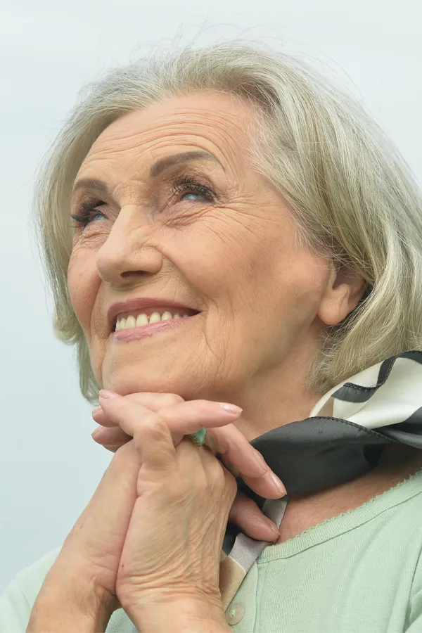 An older woman holds her hands together under her chin and looks up at a cool sky smiling. Get treatment for hot flashes and night sweats from Carolyn Zaumeyer, MSN, APRN at LowTE Florida® in Greater Fort Lauderdale.