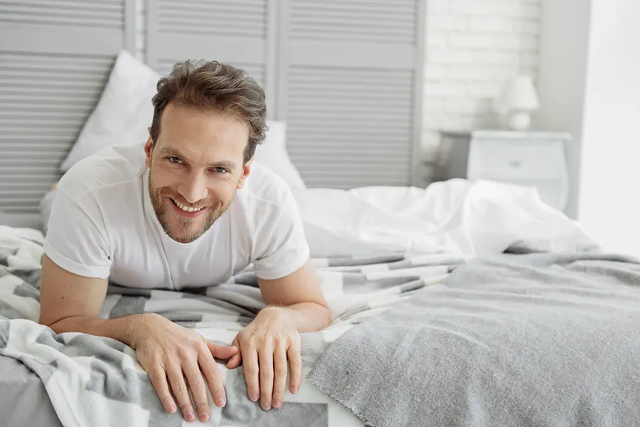 A dirty-blonde haired man lays on his stomach in bed smiling. Schedule treatment for low libido in men from Carolyn Zaumeyer, MSN, APRN at LowTE Florida® in Greater Fort Lauderdale.