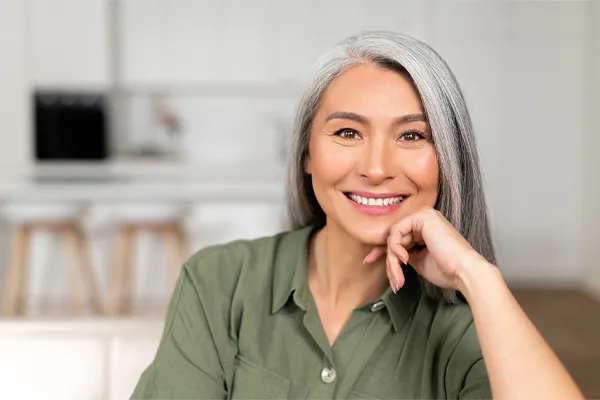 An asian woman with long gray hair and a green shirt sitting and smiling on her couch, happy with her menopause care from [NAME} in Greater Fort Lauderdale.