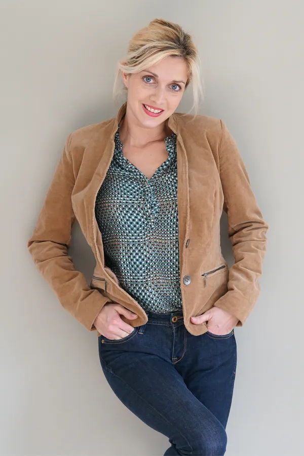 A smiling blonde woman in a tan leather jacket leans against a gray wall. Schedule metabolic syndrome from Carolyn Zaumeyer, MSN, APRN at LowTE Florida® in Greater Fort Lauderdale.