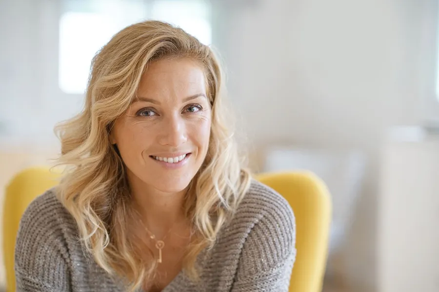 A blonde woman wearing a a gray sweater sits inside in a yellow chair, smiling and happy with her perimenopause treatment from Carolyn Zaumeyer, MSN, APRN at LowTE Florida® in Greater Fort Lauderdale.