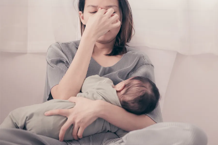 A woman nursing an infant on couch rubs her eyes with exhaustion. Get treatment for postpartum hormone imbalance from Carolyn Zaumeyer, MSN, APRN at LowTE Florida® in Greater Fort Lauderdale.