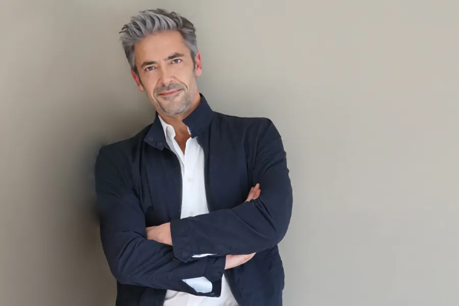 A middle-aged man in a white shirt and dark blue jacket leans against a gray wall smiling. Get testosterone for Low T from Carolyn Zaumeyer, MSN, APRN at LowTE Florida® in Greater Fort Lauderdale.